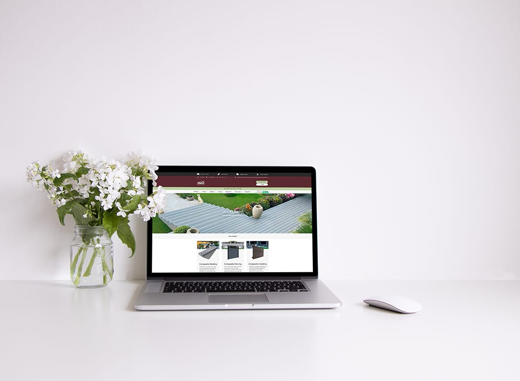 Laptop displaying a home improvement website with a focus on composite decking and fencing, accompanied by a vase of white flowers on a clean white desk.