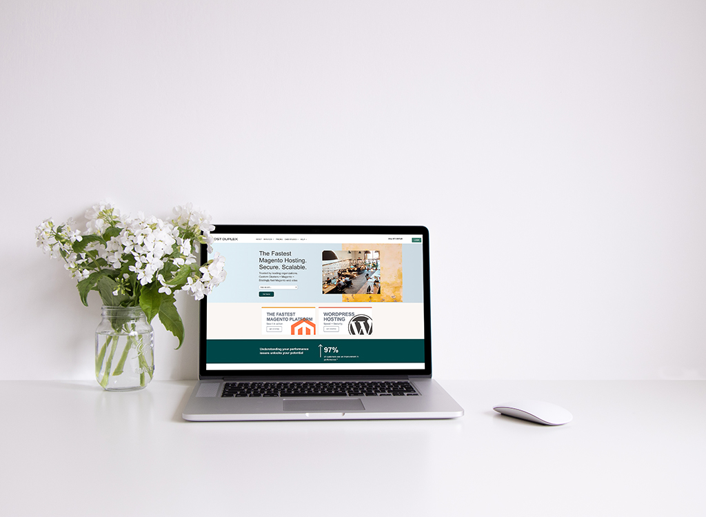 A sleek laptop displaying a website for fast and secure Magento and WordPress hosting, positioned on a minimalist desk with a vase of white flowers, showcasing a modern workspace aesthetic.