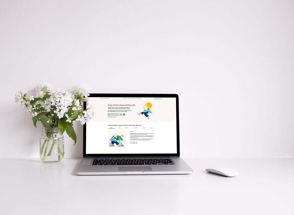 Laptop on a minimalist desk displaying a mental health website with a vibrant illustration, alongside a vase of white flowers, creating a serene workspace atmosphere.