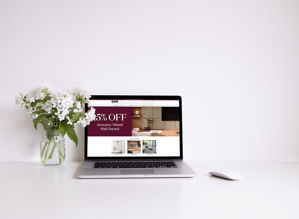 Laptop displaying a website offering 15% off on acoustic wood wall panels, accompanied by a vase of white flowers on a clean desk.