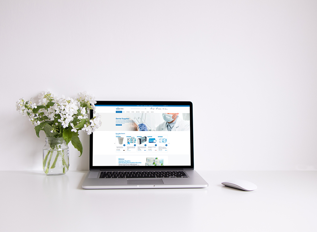 A modern laptop displaying a dental supplies website, accompanied by a vase of white flowers, on a clean white desk.