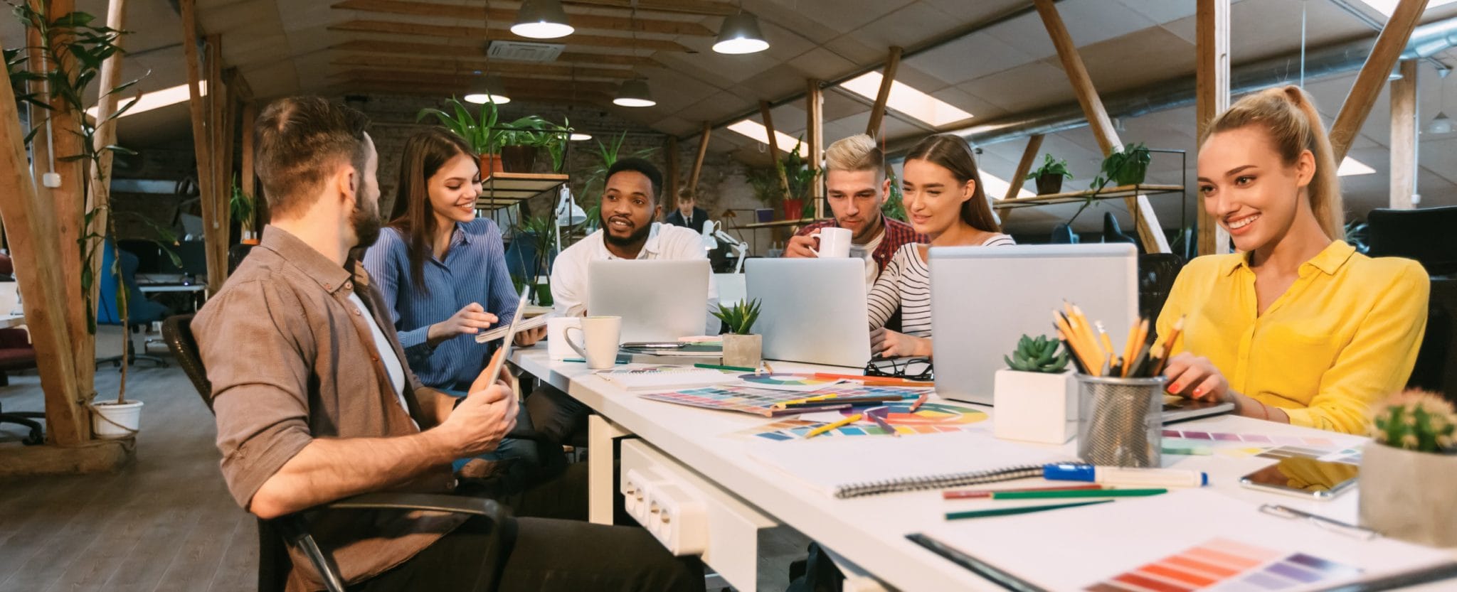 Group of diverse young professionals collaborating in a modern office space, engaged in a creative brainstorming session with laptops, tablets, and design materials on the table.